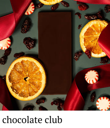 Dark chocolate bar surrounded by dried oranges, cranberries, and red ribbons on a dark surface with 'chocolate club' branding.