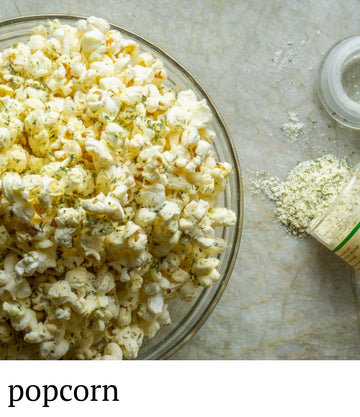 Glass bowl filled with popcorn next to a small container of seasoning on a marble surface.