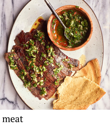 Plated dish of meat with a side of chimichurri sauce and bread on a marble surface.