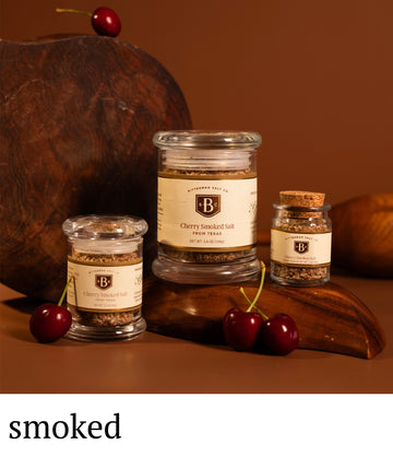 Three jars of smoked cherry salt on a wooden surface with cherries and a wooden board.