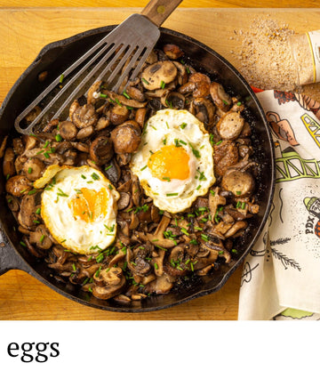 Skillet with mushrooms and eggs on a wooden surface, with a spatula and a paper towel roll in the background.