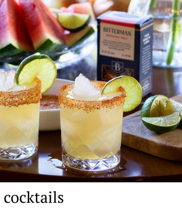 Two cocktails with lime wedges and a box of Bitterman's on a wooden surface.
