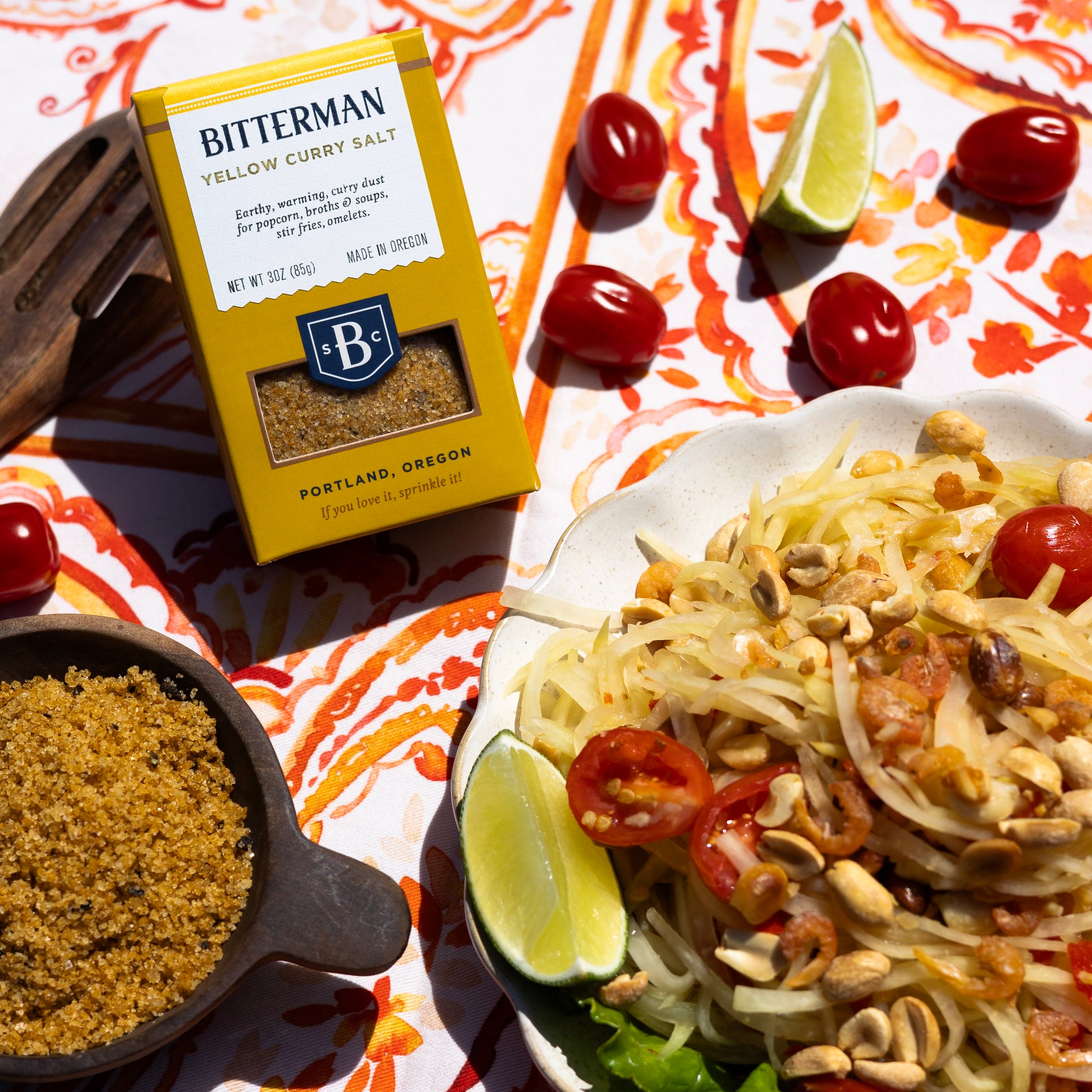 Bitterman's Yellow Curry Salt on colorful tablecloth next to noodle dish and cherry tomatoes
