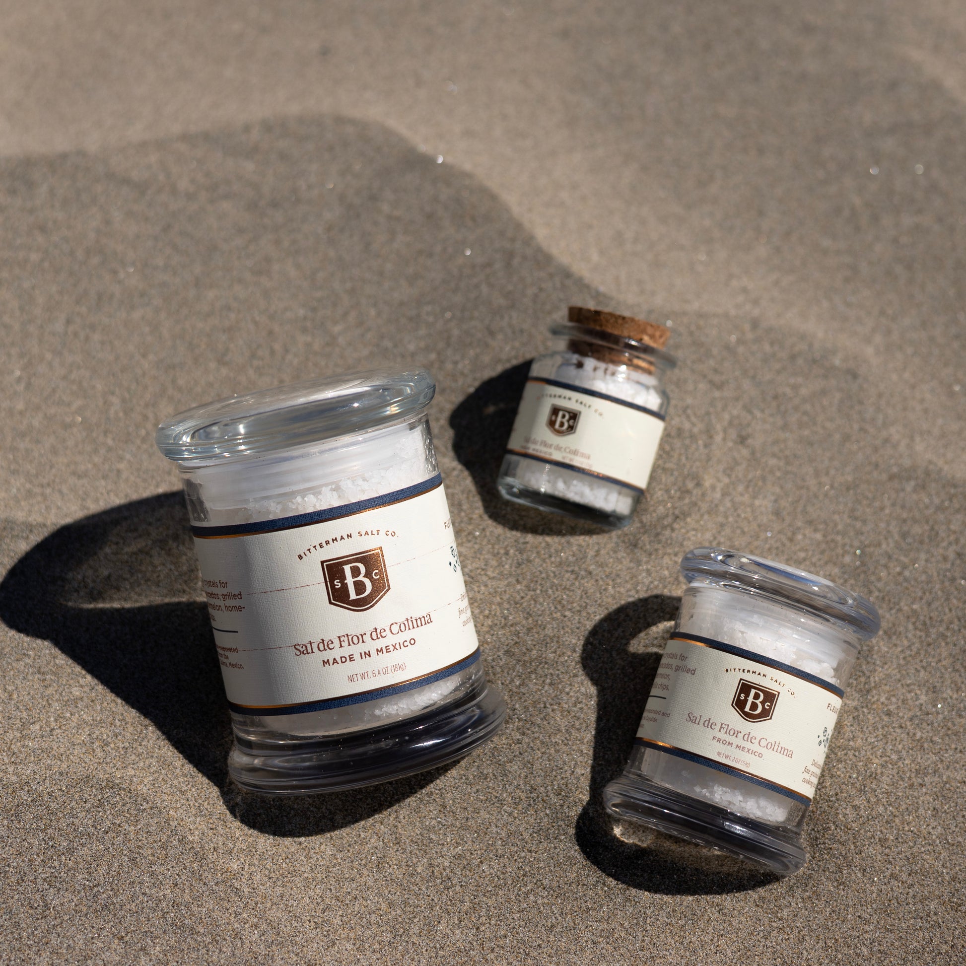 Three glass containers of 'Bitterman' brand salt on a sandy surface