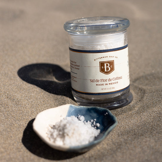 Jar of Bitterman's Salt Co. salt on sand with a small dish of salt next to it.