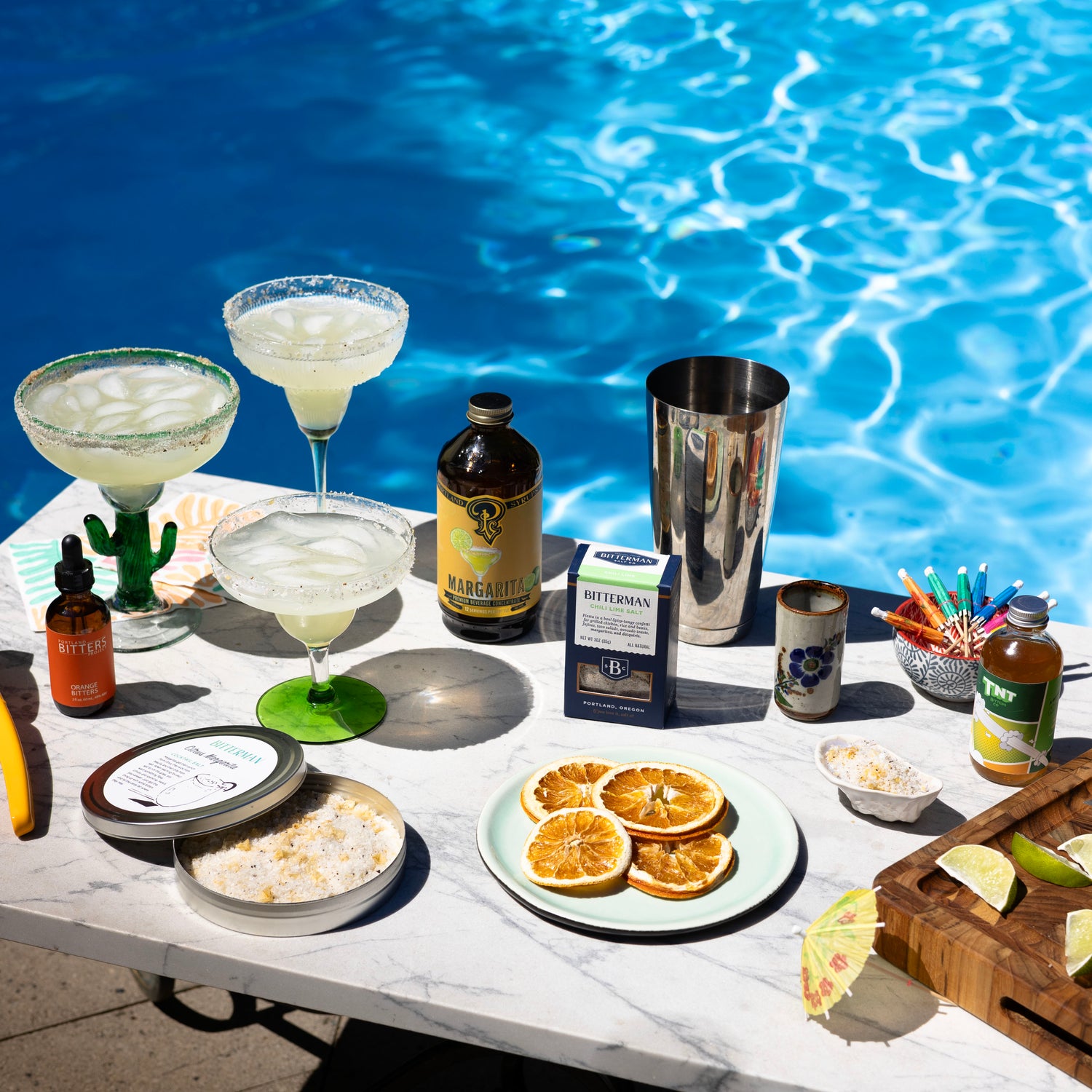 Table by a pool with drinks, garnishes, and ingredients for cocktails.