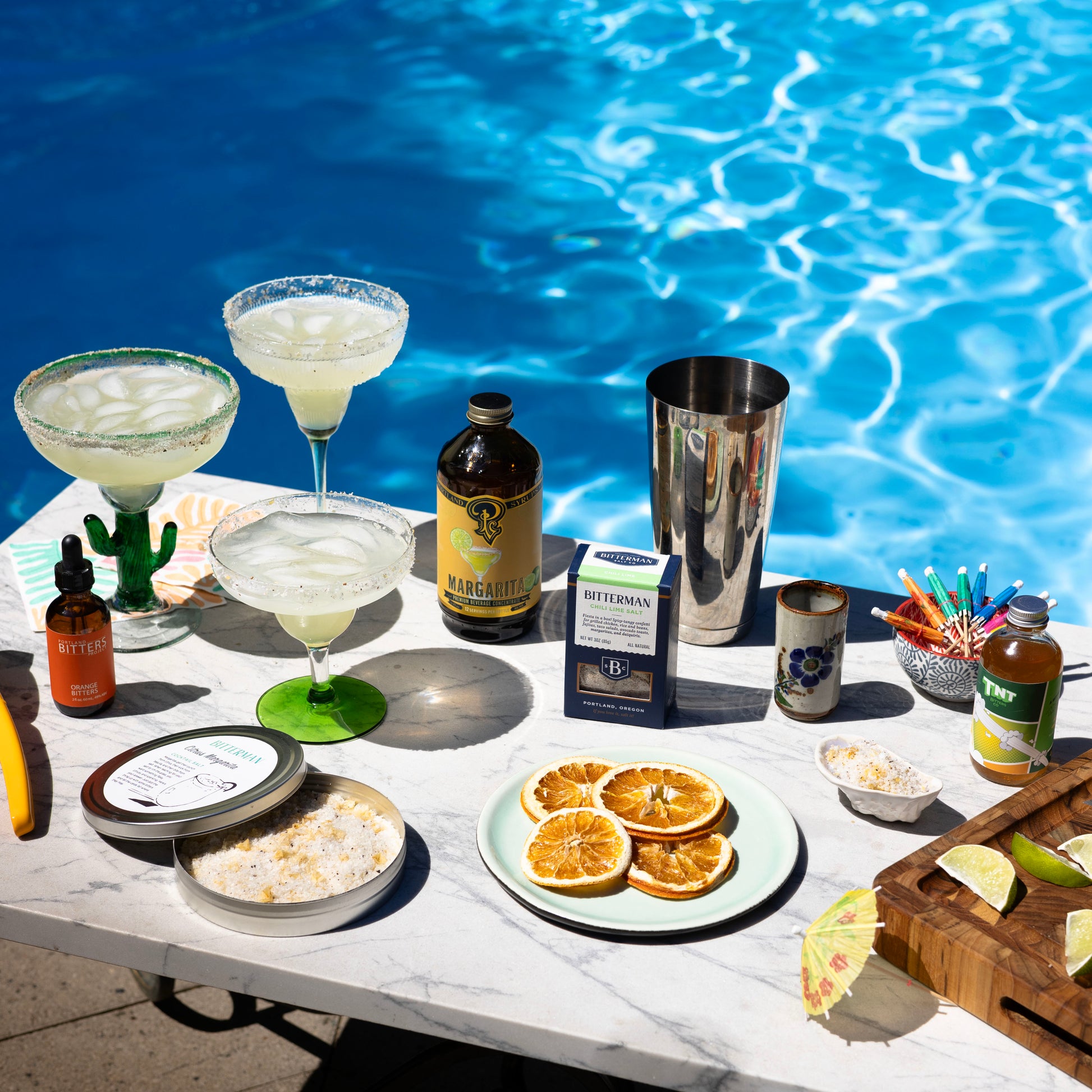Table by a pool with drinks, garnishes, and ingredients for cocktails.