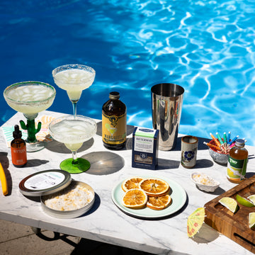 Table by a pool with drinks, garnishes, and ingredients for cocktails.