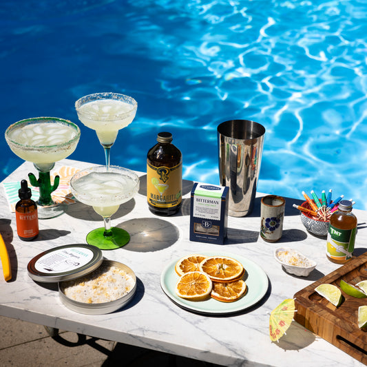 Table by a pool with drinks, garnishes, and ingredients for cocktails.