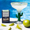 Cactus-shaped margarita glass with a drink, Bitterman's product box, and limes on a poolside table.