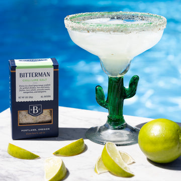 Cactus-shaped margarita glass with a drink, Bitterman's product box, and limes on a poolside table.