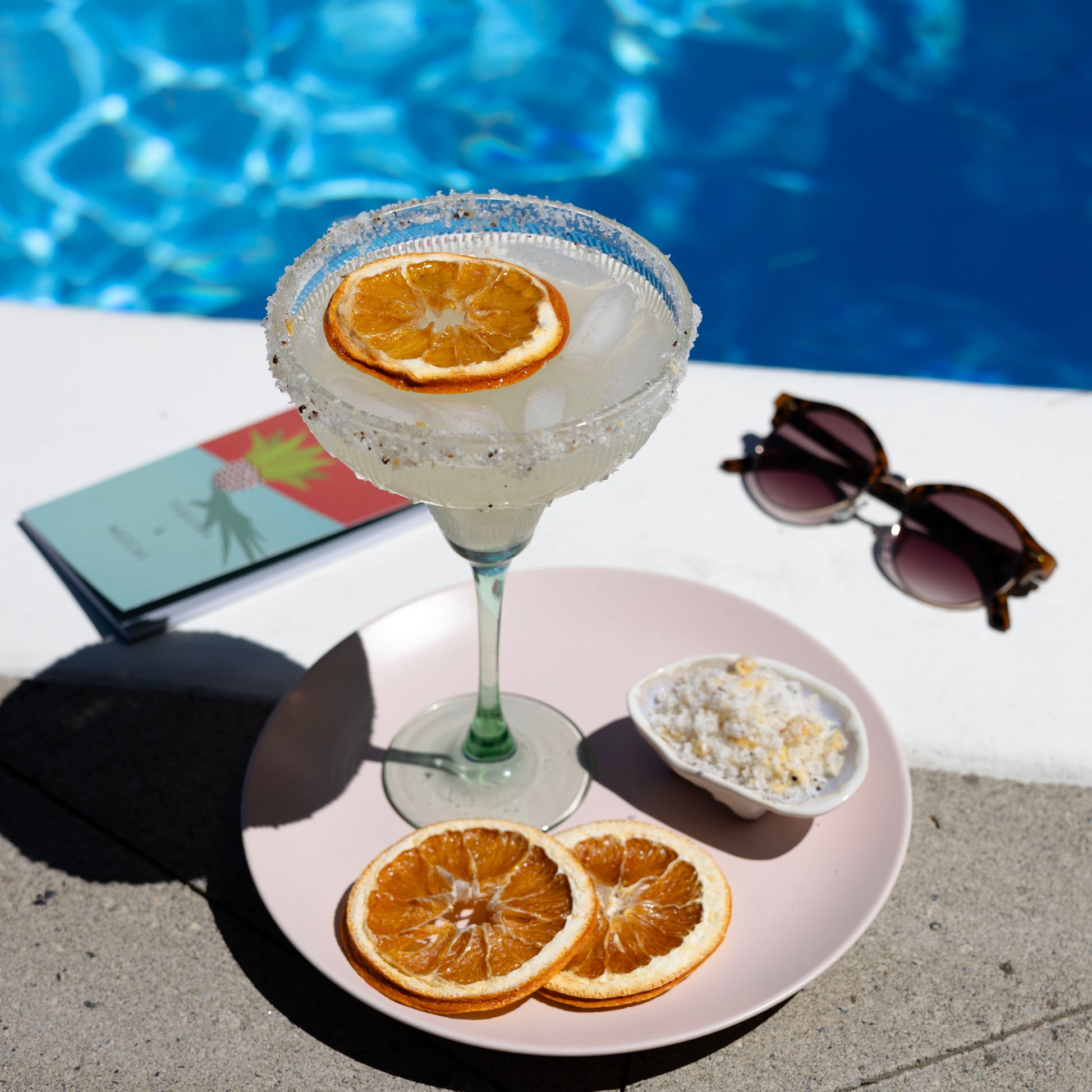 Cocktail with orange slice garnish on a table by a pool, with sunglasses and a book in the background.
