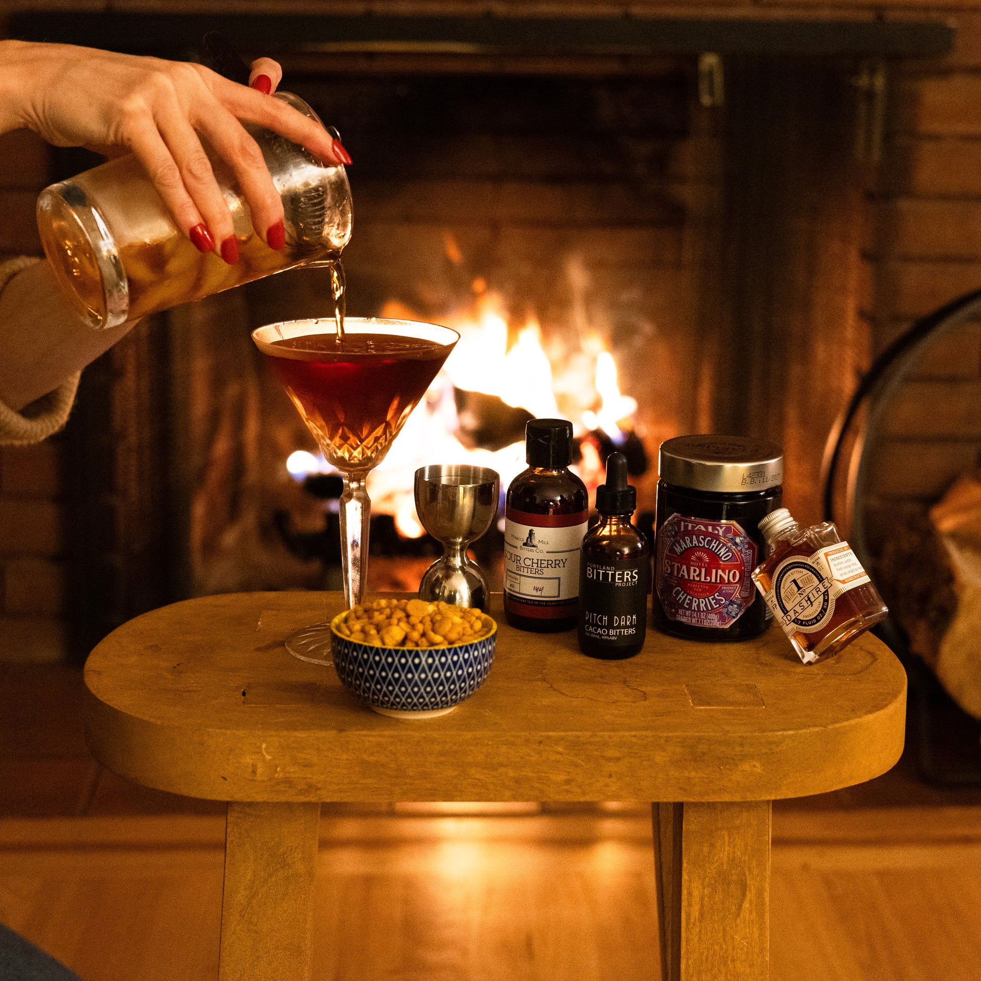 Person pouring a manhattan into a glass with a fireplace in the background cocktail club