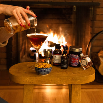 Person pouring a manhattan into a glass with a fireplace in the background cocktail club