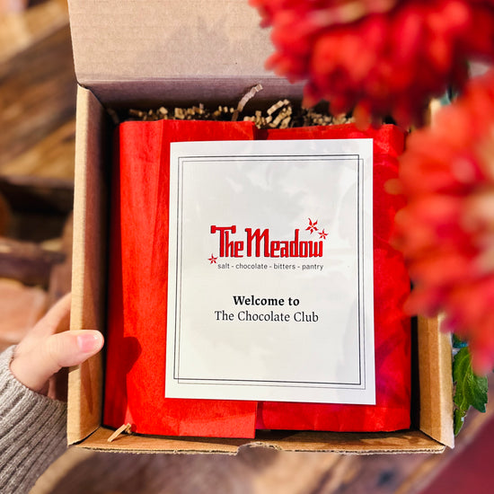 a hand holding a box with red tissue paper and a pamphlet that says welcome to the chocolate club