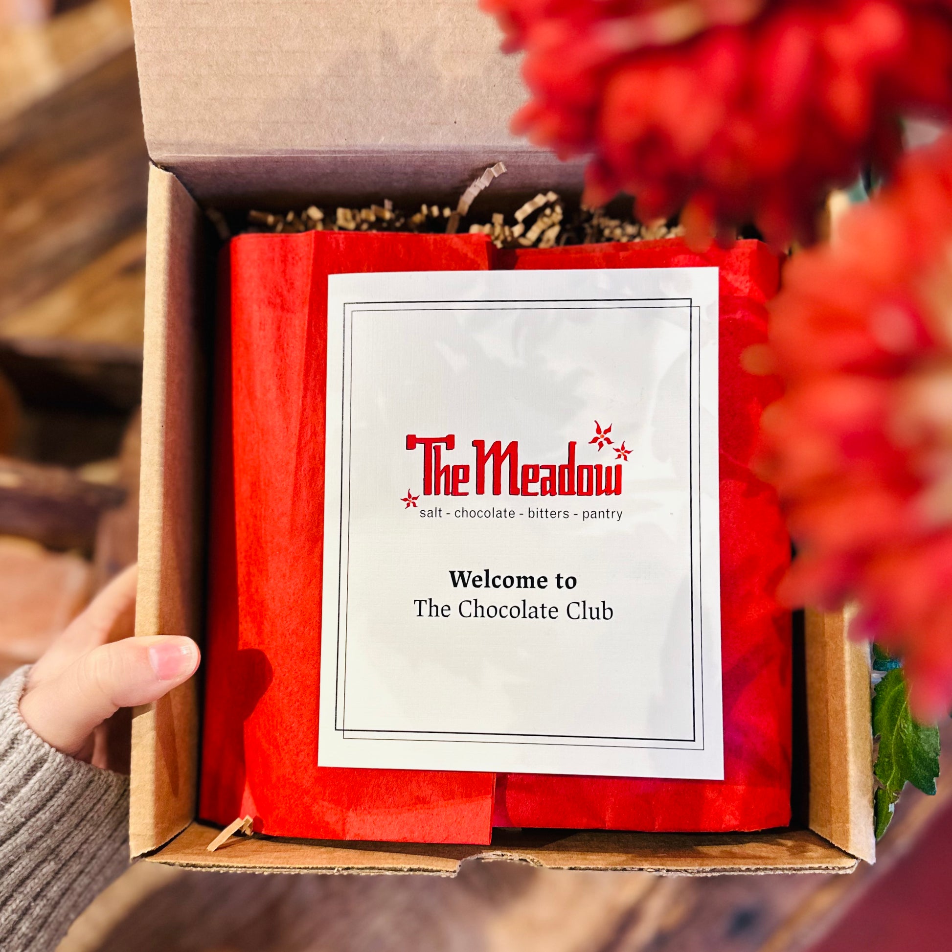 a hand holding a box with red tissue paper and a pamphlet that says welcome to the chocolate club
