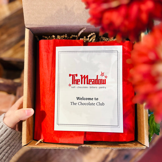 a hand holding a box with red tissue paper and a pamphlet that says welcome to the chocolate club