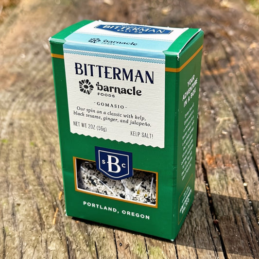 A green and white box of Bitterman x Barnacle Gomasio Salt, on a weathered wooden surface outdoors.