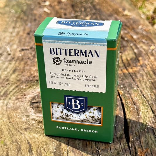 A green and white box of Bitterman x Barnacle Kelp Flake Salt, on a weathered wooden surface outdoors.
