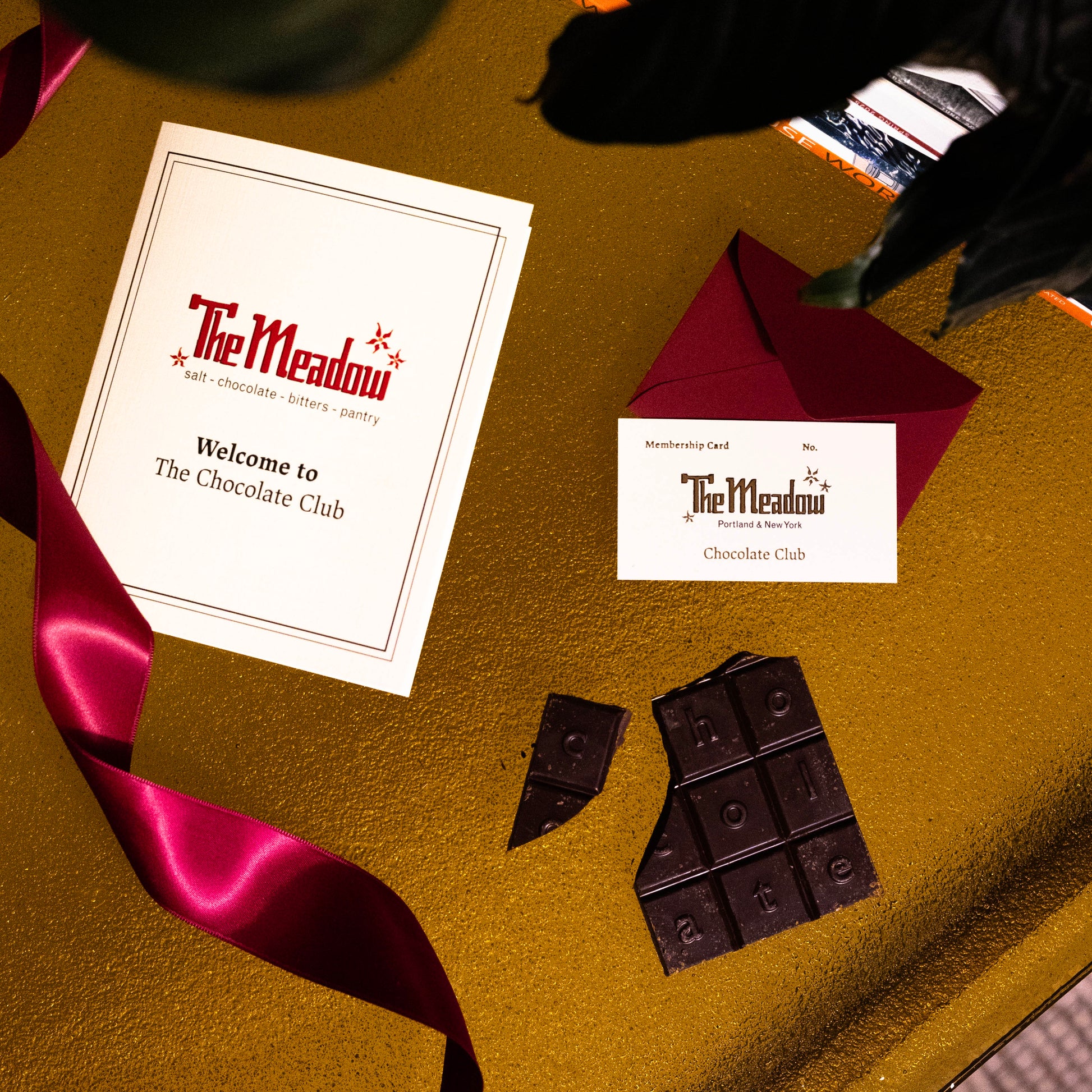 Chocolate bars and 'The Meadow' chocolate club cards on a gold surface with a red ribbon.