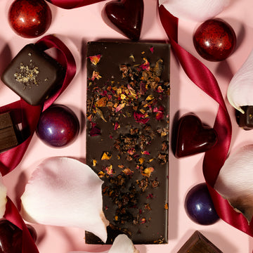 Dark chocolate bar with dried flowers surrounded by heart-shaped chocolates and ribbons on a pink background