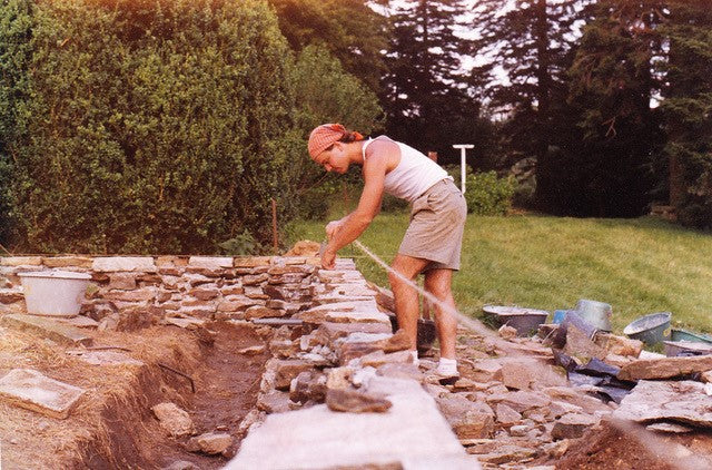 Mark Bitterman in a white tank top and khaki shorts, laying stones to build a wall